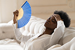 Person sweltering on couch fanning self with hand fan waiting for air conditioner repair in Ocean County
